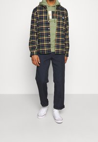 Camicia di flanella a quadri nei colori blu navy, verde e giallo, indossata sopra un hoodie verde, abbinata a pantaloni scuri e sneakers bianche.