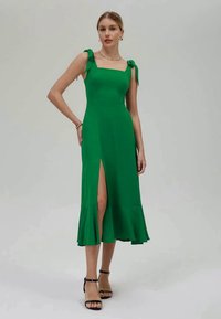 Vestido verde, sem mangas, feito de tecido suave. Apresenta decote quadrado, alças atadas nos ombros e uma abertura frontal. Bainha justa.