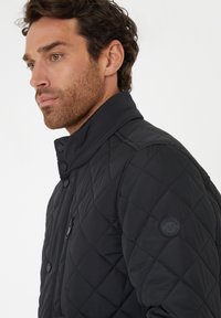 Zwarte gewatteerde jas met een hoge kraag, voorzien van een ritszak en een ronde logopatch op de schouder. Zacht materiaal met een gladde textuur.