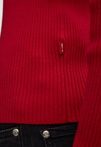 Czerwony sweter prążkowany z małą czerwoną metką z białym wykrzyknikiem. Teksturowany wzór dzianiny, dopasowany fason, w połączeniu z czarnymi dżinsami.