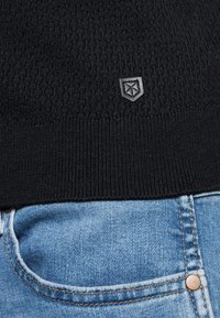 Svart stickad tröja med ett texturerat mönster och ribbad fåll, accentuerad med en liten grå emblem. Matchad med blå denimjeans.
