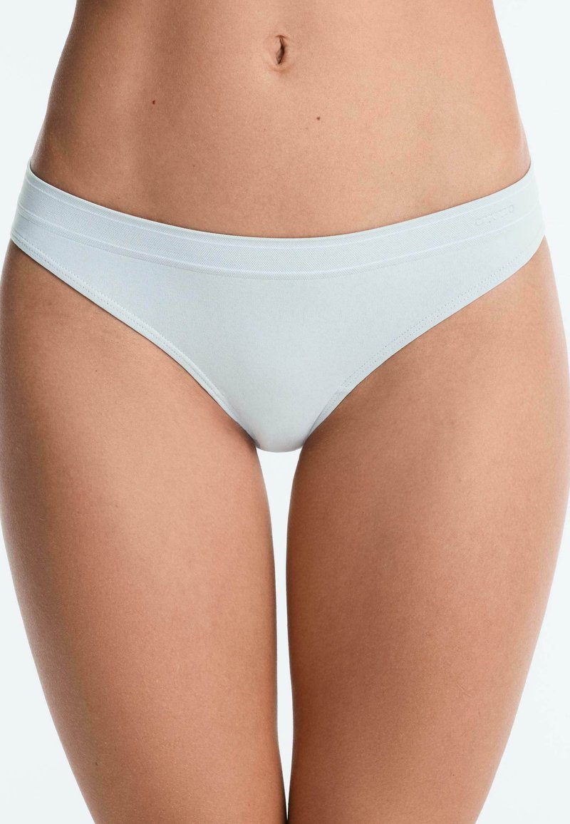 Femme portant un sous-vêtement blanc uni, sans coutures, style bikini, montrant le torse de la taille jusqu'à mi-cuisse sur un fond neutre.