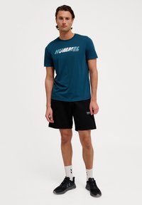 Tealfärgad t-shirt med kort ärm och "HUMMEL"-logotyp, tillsammans med svarta shorts. Svarta sneakers och vita strumpor med ett zigzag-mönster fulländar looken.