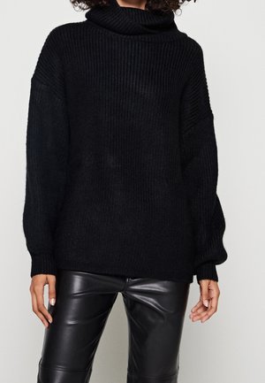 Femme portant un pull à col roulé côtelé noir oversize et un pantalon en cuir noir, debout devant un fond clair uni.
