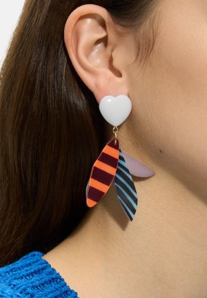 Zbliżenie ucha noszącego biały kolczyk w kształcie serca z trzema zwisającymi, paskowanymi zawieszkami w kształcie liści w kolorach pomarańczowym, niebieskim i fioletowym.