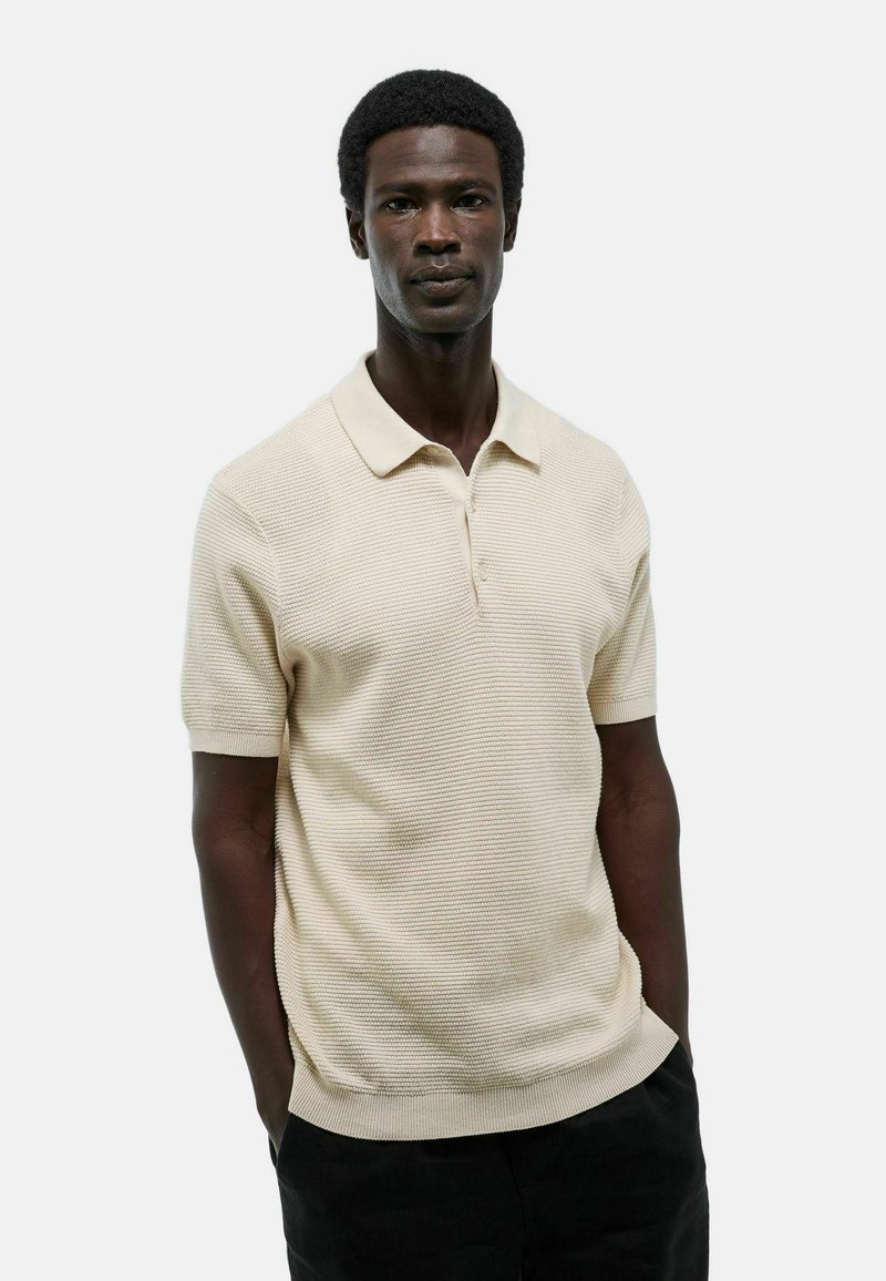 Polo shirt beige à manches courtes avec un motif texturé, patte de boutonnage à trois boutons et col et ourlet côtelés. Porté avec un pantalon noir.