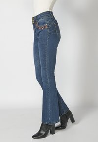 Flared blue jeans with a high waist, embellished front pockets, and gold buttons. Paired with black heeled ankle boots.