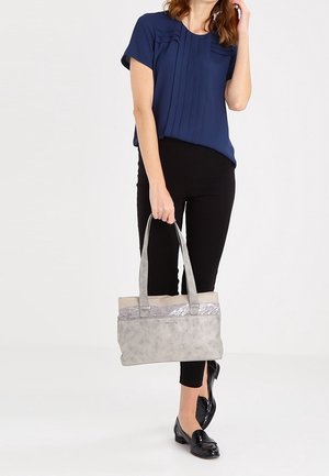 Donna in blusa blu navy e pantaloni neri che tiene una borsa grigia con manici lunghi, in piedi su uno sfondo chiaro e semplice.