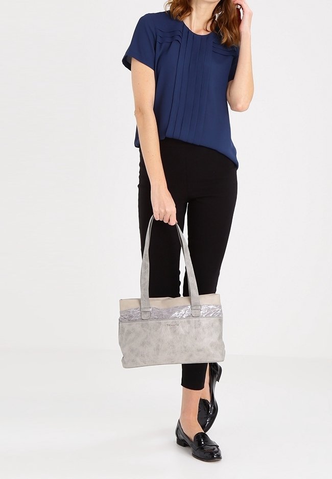 Donna in blusa blu navy e pantaloni neri che tiene una borsa grigia con manici lunghi, in piedi su uno sfondo chiaro e semplice.