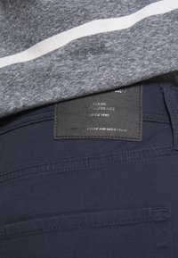 Námořnické modré džíny s hladkou texturou, doplněné černou koženou nášivkou s nápisem "Jeans Intelligence" a pokyny k údržbě.