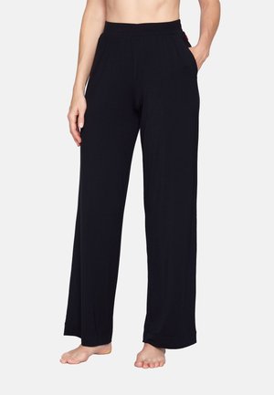 Personne portant un pantalon large noir et ample avec des poches, debout pieds nus contre un fond clair uni.