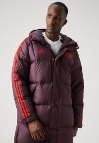 Abrigo largo acolchado morado con capucha, presenta franjas rojas de Adidas en las mangas y un logo en el pecho, textura suave, cierre de broche y cremallera.