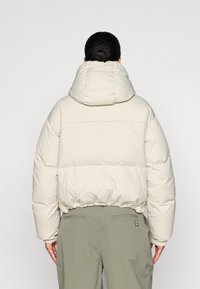 Zachte beige jas met capuchon, elastische onderrand en gestikte panelen. Draag je over olijfgroene broeken. Eenvoudig, functioneel ontwerp.