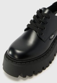 Black patent leather shoe with a chunky rubber sole. Features flat black laces, rounded toe, and textured platform with jagged edges.