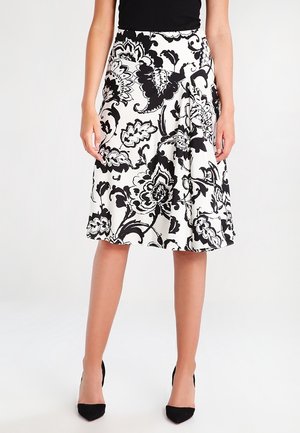 Woman wearing a knee-length white skirt with large black floral patterns, paired with black pointed-toe heels and a black sleeveless top.