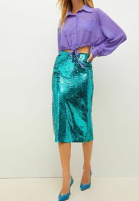 Blusa corta viola con due tasche frontali; gonna midi turchese con paillettes e forma aderente; abbinata a tacchi alti coordinati.