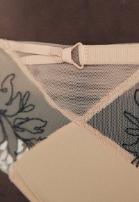 Beige lingerie featuring a mesh design with black floral embroidery, an adjustable strap, and smooth fabric detailing.