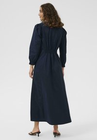 Navyblaues Baumwollkleid mit langen Puffärmeln, gerafftem Bund und fließender Silhouette, die bis zu den Knöcheln reicht. Getragen mit schwarzen hohen Sandalen.
