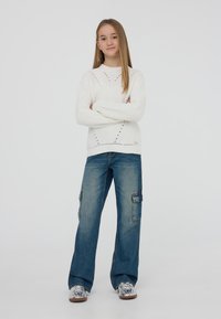 Biały sweter z dzianiny o strukturalnym wzorze, zestawiony z luźnymi niebieskimi spodniami cargo z bocznymi kieszeniami i casualowymi trampkami w wzory.