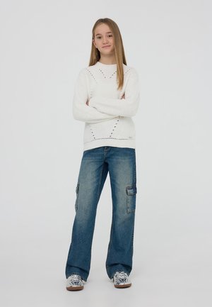 Biały sweter z dzianiny o strukturalnym wzorze, zestawiony z luźnymi niebieskimi spodniami cargo z bocznymi kieszeniami i casualowymi trampkami w wzory.