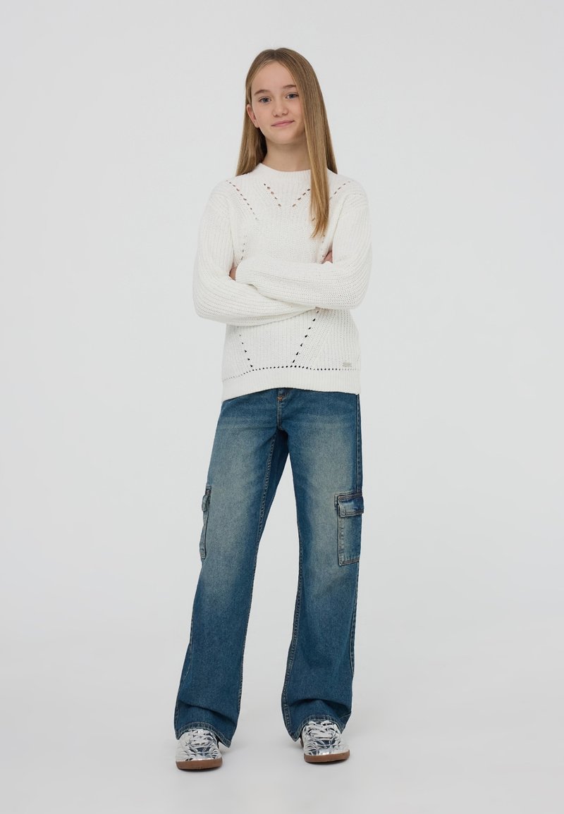 Biały sweter z dzianiny o strukturalnym wzorze, zestawiony z luźnymi niebieskimi spodniami cargo z bocznymi kieszeniami i casualowymi trampkami w wzory.