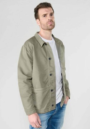 Light olive green jacket with a button-up design, two front pockets, and a collar. Worn over a white t-shirt and paired with denim jeans.