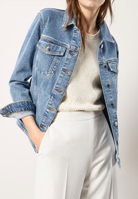 Nainen, jolla on vaaleansininen farkkutakki vaalean kerrotun villaneuleen ja korkeavyötäröisten valkoisten housujen päällä, käsi taskussa, yksinkertaista taustaa vasten.