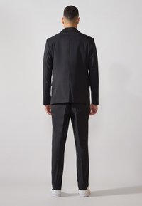 Sacou negru cu spate drept, asociat cu pantaloni asortați. Prezintă un material neted și detalii fine de cusătură, purtat cu teniși albi.