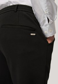 Zwarte sweatpants hebben een gladde textuur, een elastische tailleband en een klein zilveren label met de tekst "JP 1980." Bevat een detail van een zijzak.
