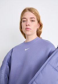Sudadera púrpura con cuello ajustado, que presenta un logo de Nike en blanco. El material parece suave, con costuras visibles y una textura lisa.
