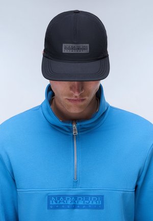 Man draagt een zwarte Napapijri pet en een blauwe zip-up trui met het Napapijri-logo op de borst, kijkt omlaag tegen een effen achtergrond.