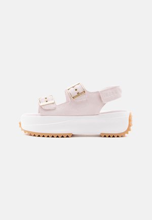 Sandale plateforme rose pâle avec deux boucles dorées, semelle blanche épaisse, semelle extérieure marron dentelée et bride de talon ouverte sur fond blanc.