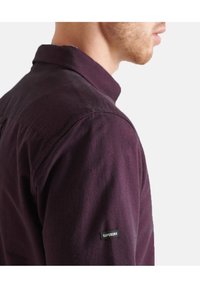 Uomo che indossa una maglietta a maniche lunghe di un viola scuro con un'etichetta Superdry sulla manica superiore, mostrato in profilo laterale su sfondo bianco.