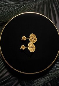 Guldtonade stiftörhängen med texturerad blommönsterdesign. Uppställda på en cirkulär svart displayplatta, omgivna av gröna växtblad.