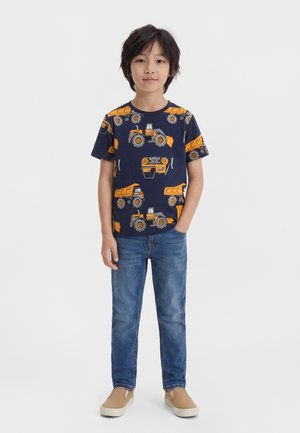 Chłopiec stoi, ubrany w granatowy t-shirt z pomarańczowymi pojazdami budowlanymi, niebieskie dżinsy i jasnobrązowe buty wsuwane na białym tle.