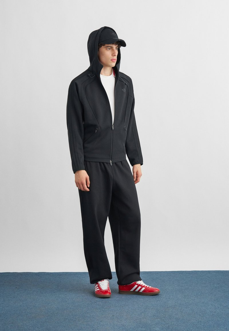 Junger Mann steht und trägt schwarzen Hoodie, schwarze Mütze, schwarze Hose, weißes Hemd und rote Adidas-Sneaker auf einem blau beflockten Boden.