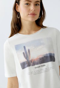 Weißes Baumwoll-T-Shirt mit einem Sonnenuntergang-Print, der einen Kaktus zeigt. Unten steht der Text "Sonnenuntergangspfad" mit poetischen Phrasen darunter.