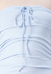 Šviesiai mėlyna be petnešėlių viršūnė su siaurais horizontaliais baltais ruožais. Turi surinktos medžiagos detalių ir raištelį nugaroje. Lygus paviršius.