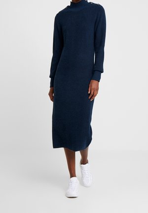 Vrouw draagt een marineblauwe midi-jurk met lange mouwen en knoopdetails op de schouders en witte sneakers, staand tegen een effen achtergrond.