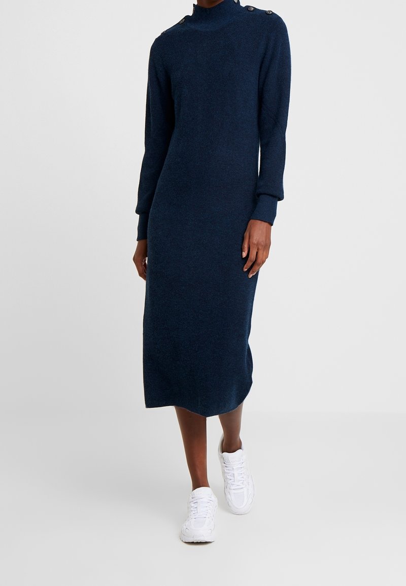 Femme portant une robe midi bleu marine à manches longues avec des boutons aux épaules et des baskets blanches, debout sur un fond uni.