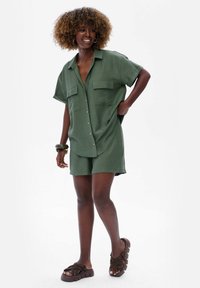 Zielona koszula z krótkim rękawem i dwiema kieszeniami na piersi, w połączeniu z dopasowanymi szortami, wykonana z lekkiego materiału. Noszona z ciemnobrązowymi sandałami.