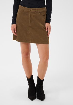 Femme portant une jupe en velours côtelé marron, un haut noir à manches longues et des bottines noires, debout devant un fond uni.