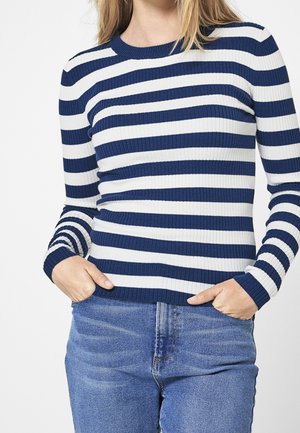 Vrouw in een blauw-wit gestreepte trui met lange mouwen en een blauwe spijkerbroek, met haar handen in de zakken, cropped tot boven de schouders.