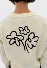 Pull crème avec des motifs abstraits de trois fleurs en noir dans le dos, porté par une personne aux cheveux bruns courts de dos.