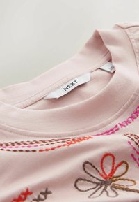 Jasnoróżowy dekolt koszuli z haftowanymi, wielokolorowymi wzorami kwiatowymi i ściegami, z białą metką marki z napisem "NEXT".