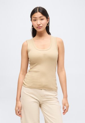 Mladá žena s dlhými tmavými vlasmi nosí zlaté kruhové náušnice, béžový rebrovaný tielko a svetlé nohavice na jednoduchom pozadí.