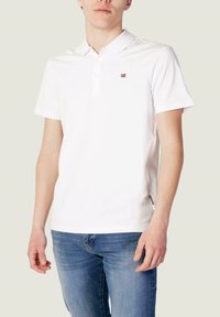 Polo shirt blanc à manches courtes, avec une patte de boutonnage et un petit emblème rouge sur la poitrine. Le tissu semble lisse avec une coupe classique.