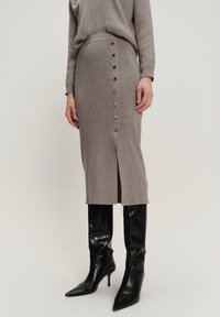 Vrouw in een taupe ribgebreide rok met zijknopen en een split, gecombineerd met een bijpassende top en zwarte puntschoen kniehoge laarzen.