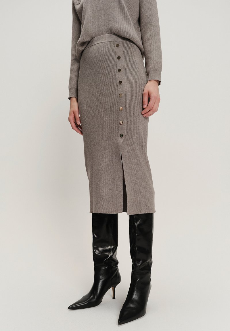 Vrouw in een taupe ribgebreide rok met zijknopen en een split, gecombineerd met een bijpassende top en zwarte puntschoen kniehoge laarzen.