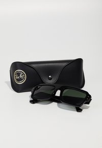 Ochelari de soare Ray-Ban negri cu lentile verzi închise, având un cadru pătrat și accente aurii. Împreună cu un toc din piele texturată neagră.